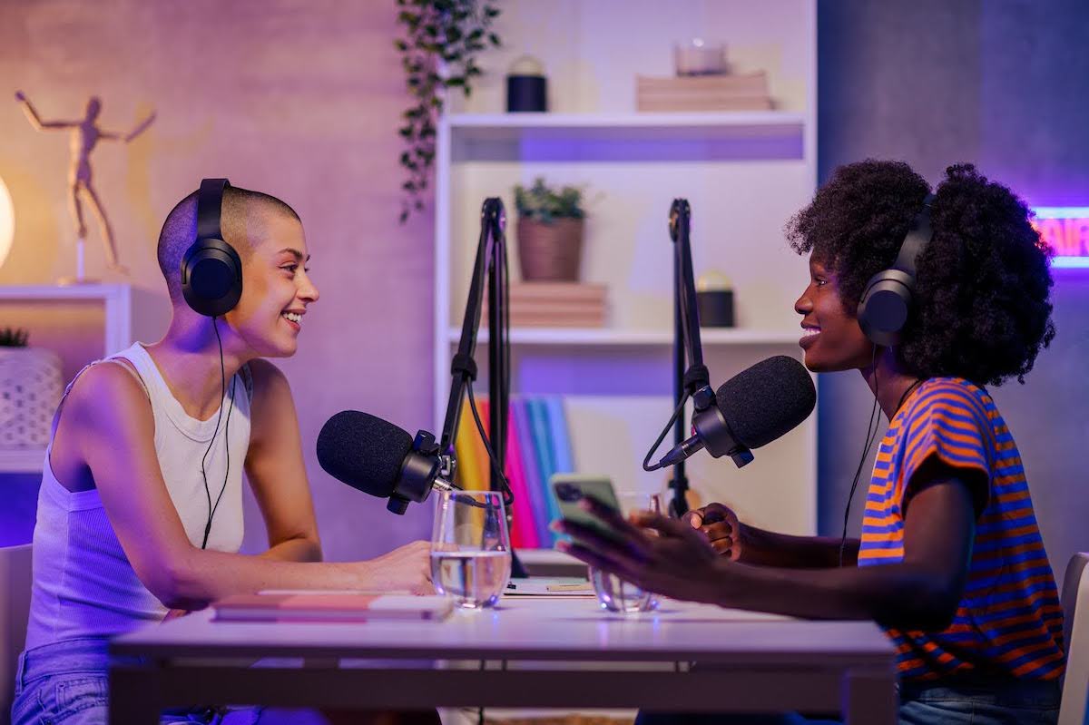 Two podcasters speaking and laughing into microphones at a home studio